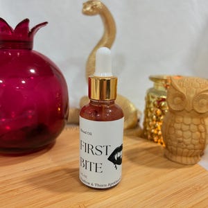 May include: A small glass dropper bottle containing a reddish liquid with a white label that reads Ritual Oil FIRST BITE 1 FL OZ Shadow & Thorn Apothecary and features a graphic illustration of a mouth with fangs and a drop of blood.