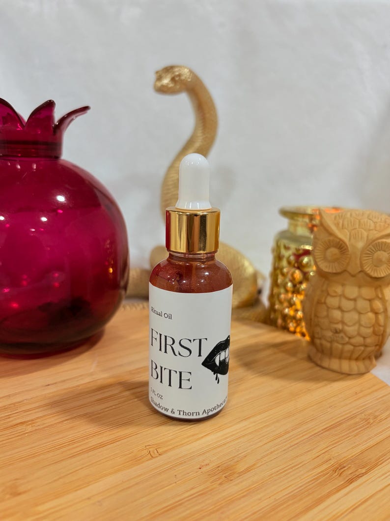 May include: A small glass dropper bottle containing a reddish liquid with a white label that reads Ritual Oil FIRST BITE 1 FL OZ Shadow & Thorn Apothecary and features a graphic illustration of a mouth with fangs and a drop of blood.
