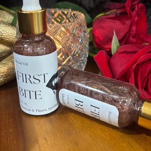 May include: Two glass dropper bottles containing a dark, shimmering liquid labeled Ritual Oil FIRST BITE by Shadow & Thorn Apothecary. Each bottle has a gold collar, a white dropper top, and a 1 FL OZ capacity. One bottle stands upright while the other lies on its side.