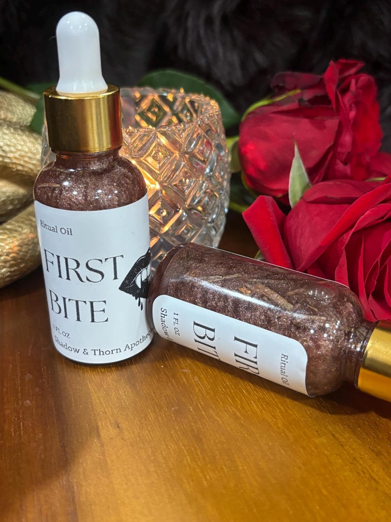 May include: Two glass dropper bottles containing a dark, shimmering liquid labeled Ritual Oil FIRST BITE by Shadow & Thorn Apothecary. Each bottle has a gold collar, a white dropper top, and a 1 FL OZ capacity. One bottle stands upright while the other lies on its side.