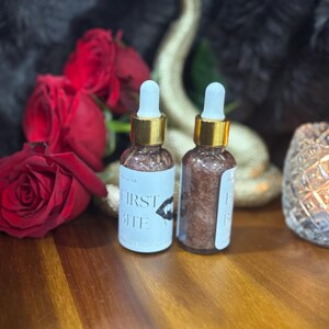 May include: Two clear glass dropper bottles filled with a shimmering bronze liquid. Each bottle features a white label with the text "FIRST BITE" and a graphic of lips. The bottles have gold-colored collars and white dropper tips.