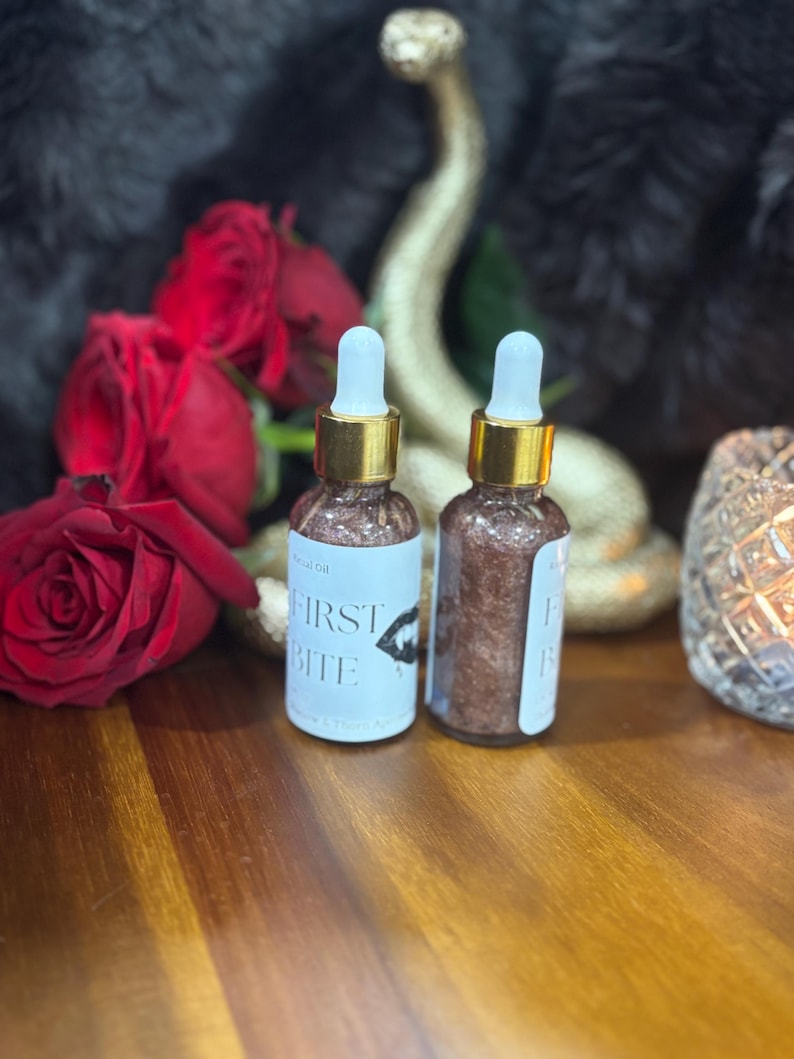 May include: Two clear glass dropper bottles filled with a shimmering bronze liquid. Each bottle features a white label with the text "FIRST BITE" and a graphic of lips. The bottles have gold-colored collars and white dropper tips.