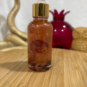 May include: A clear glass dropper bottle filled with a shimmering, copper-toned liquid containing a single visible rose petal and small dark particles at the base. The bottle features a metallic gold collar and a white rubber dropper top.