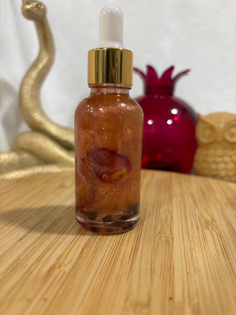May include: A clear glass dropper bottle filled with a shimmering, copper-toned liquid containing a single visible rose petal and small dark particles at the base. The bottle features a metallic gold collar and a white rubber dropper top.