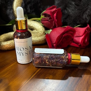 May include: Two glass bottles with gold-colored caps and white droppers. One bottle is labeled "Sacred Ritual Oil ROSA MYSTICA" and the other is labeled "Red Thread Collection". Red roses and a gold snake are in the background.