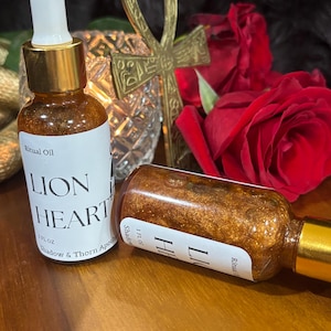 May include: Two bottles of "Lion Heart" ritual oil with gold accents and white labels. The bottles are filled with a shimmering, golden liquid. An Ankh and red roses are in the background. The label also reads "Shadow & Thorn Apothecary".