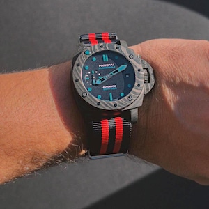 Peut inclure: Une montre automatique Panerai Submersible avec un boîtier et une lunette en fibre de carbone marbrée gris foncé. Le cadran présente des index et des aiguilles luminescents bleu clair, un petit cadran des secondes et un guichet de date.