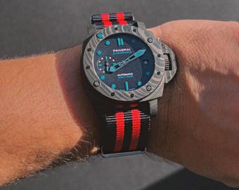 May include: A Panerai Submersible automatic watch with a dark gray marbled carbon fiber case and bezel. The dial features light blue luminous markers and hands, a small seconds sub-dial, and a date window. It has a black and red striped fabric strap.
