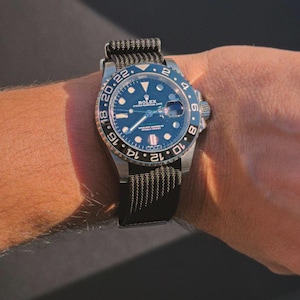 Op de afbeelding: Een Rolex GMT-Master II horloge van roestvrij staal met een blauwe wijzerplaat en een zwarte en olijfgroene stoffen band. Het horloge heeft een draaibare bezel met zilveren cijfers en een datumweergave. Het horloge zit om een pols.