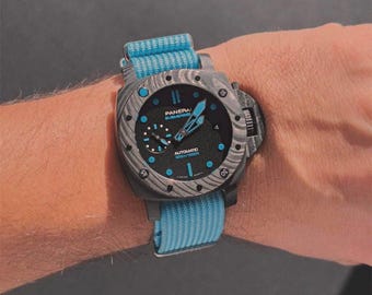 May include: A Panerai Submersible watch with a black dial, bright blue accents, and a textured grey case. The watch features a black face with the text Panerai Submersible and Automatic 300m/1000ft, paired with a bright blue fabric strap.