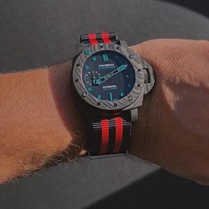 Könnte beinhalten: Eine Luxus-Armbanduhr mit einem schwarz-rot gestreiften Armband. Das Zifferblatt der Uhr ist dunkelblau mit türkisfarbenen Akzenten und die Marke "Panerai" ist sichtbar. Die Uhr befindet sich an einem Handgelenk.