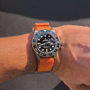 Può includere: Un orologio Rolex in acciaio inossidabile con quadrante nero e lunetta blu e nera. È fissato con un cinturino in nylon arancione brillante. Il quadrante mostra il logo Rolex e indicatori temporali.