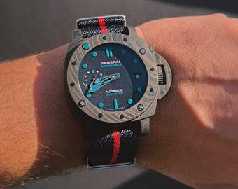 May include: A Panerai Submersible watch with a dark blue face and a black strap with red stripes. The watch has a textured, gray bezel and a crown protector. The watch face has teal accents and the text "AUTOMATIC 300M/1000FT".