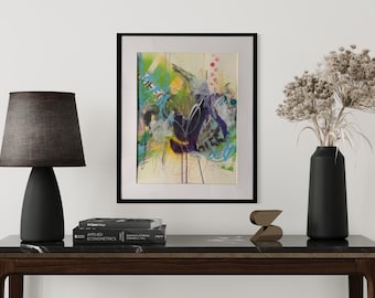 May include: A framed abstract painting featuring vibrant splashes of green, blue, and purple paint with thin black lines. The artwork is displayed in a black frame, centered above a dark console table with a black lamp, books, and a vase of dried flowers.