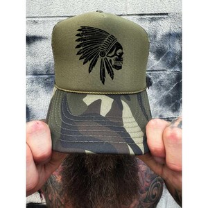 May include: Olive green trucker hat with a camouflage pattern on the brim. The hat features a black graphic of a Native American headdress and skull. The hat is being held up by a person.
