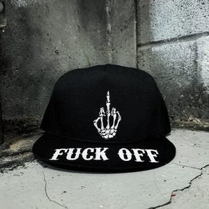 May include: Black baseball cap featuring a white skeleton hand giving the middle finger. The brim of the cap displays the words "FUCK OFF" in bold white letters. The cap is set against a textured gray background.