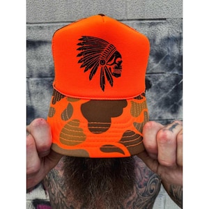 May include: An orange trucker hat with a brown camouflage pattern on the brim. The hat features a black graphic of a Native American headdress and skull on the front. The hat is being held up by a person.