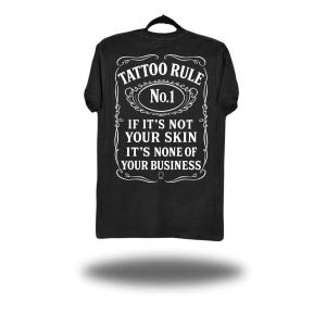 May include: Black t-shirt with white text that reads "TATTOO RULE No.1 IF IT'S NOT YOUR SKIN IT'S NONE OF YOUR BUSINESS". The text is in a decorative frame, resembling a label.