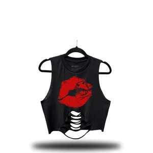 May include: Black crop top with a distressed design and a red lip graphic. The sleeveless shirt is hanging on a black hanger. The graphic features a pair of lips with a devil horn design.
