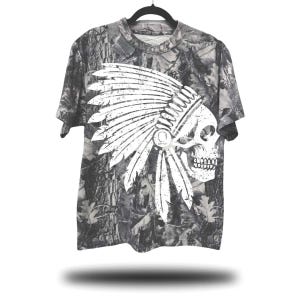May include: A gray camouflage t-shirt featuring a white skull wearing a Native American headdress. The skull graphic is distressed, giving it a vintage look. The shirt is hanging on a black hanger against a white background.
