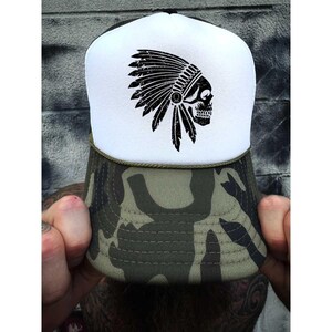 May include: A trucker hat with a white front panel featuring a black skull wearing a Native American headdress. The brim and lower portion of the hat are camouflage, with a black mesh back.