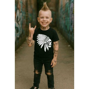 May include: A young child with a mohawk hairstyle and skull tattoos is wearing a black t-shirt with a white Native American headdress and skull graphic. The child is also wearing black ripped jeans.