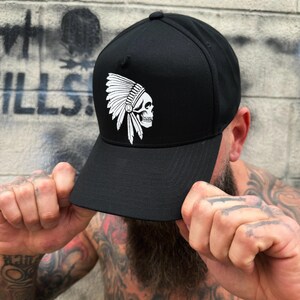 May include: Black baseball cap featuring a white graphic of a skull wearing a Native American headdress. The cap has a structured crown and a curved brim. The design is centered on the front of the cap.