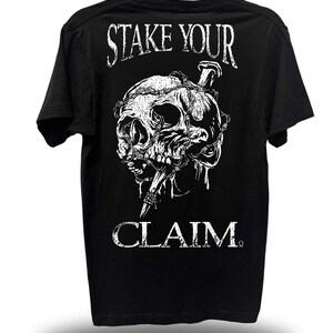 May include: Black t-shirt with a white graphic of a skull pierced by a nail and wrapped in barbed wire. The words "STAKE YOUR" are above the skull, and "CLAIM" is below.