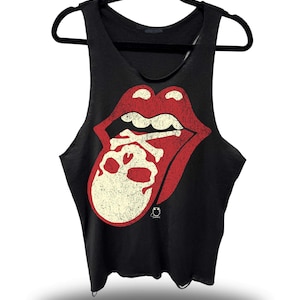 May include: Black tank top with a distressed red and cream-colored graphic of a mouth, skull, and crossbones. The sleeveless shirt is hanging on a black hanger. The graphic is a popular rock and roll design.