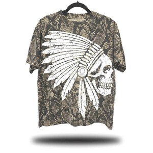 May include: A short-sleeved t-shirt with a snake skin pattern. The shirt features a distressed white graphic of a skull wearing a Native American headdress. The shirt is hanging on a black hanger against a white background.