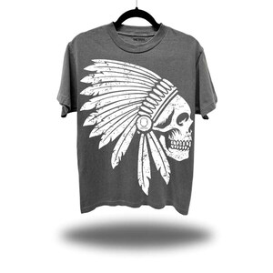 May include: Dark gray t-shirt featuring a white graphic of a skull wearing a Native American headdress. The shirt is hanging on a black hanger against a white background. The text "THE DRIVE" is visible on the shirt.