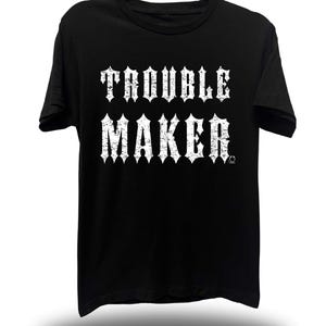 May include: Black t-shirt with the words "TROUBLE MAKER" in large, distressed white lettering. The shirt is hanging on a black hanger. The text is in a bold, gothic font.
