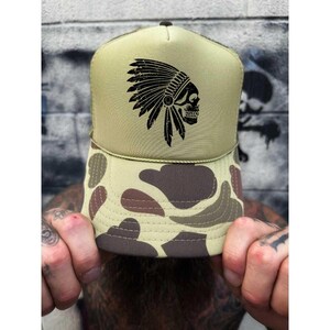May include: A trucker hat with a tan crown and a camouflage pattern on the brim. The hat features a black graphic of a Native American headdress and skull. The hat is a stylish accessory.