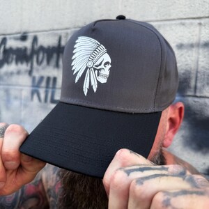 May include: A two-tone baseball cap with a black brim and a gray crown. The cap features a white graphic of a skull wearing a Native American headdress. The cap is being held by a person.