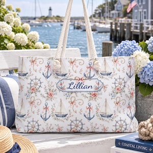 May include: A white tote bag with rope handles featuring a repeating pattern of sailboats, blue anchors, ship wheels, pink roses, and ribbons. The center of the bag displays a rectangular label with the name Lillian printed in blue script text.