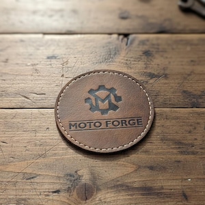 Puede incluir: Un posavasos de cuero marrón con el texto "MOTO FORGE" y un logotipo de engranaje. El posavasos es redondo con costuras blancas alrededor del borde. Sobre una superficie de madera.