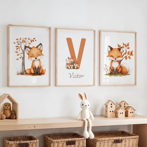 Op de afbeelding: Drie ingelijste aquarelprints met een schattige vos met oranje en bruine vacht. De prints zitten in houten frames en bevatten een boom, de letter "V" met de naam "Victor" en een vos met herfstbladeren. Een houten plank bevat een teddybeer, een konijn en houten huisjes.