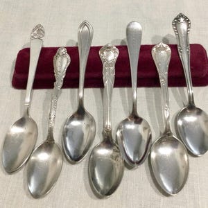 May include: Seven antique silver spoons with ornate handle designs are displayed on a burgundy velvet surface. The spoons vary in style, with detailed patterns and engraved elements. The spoons are arranged on a light-colored fabric background.