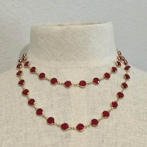 May include: A double-strand necklace featuring round, faceted red stones set in gold-toned metal bezels. The stones are evenly spaced along a delicate gold-toned chain, creating a layered look that rests against the neck.