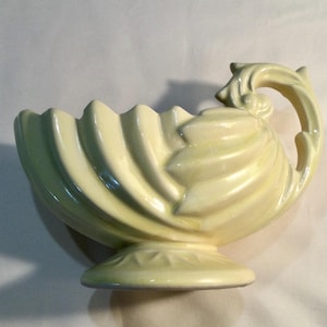 May include: A pale yellow ceramic bowl with a decorative handle. The bowl has a scalloped rim and sits on a pedestal base. The handle is curved and features a stylized floral design. The bowl is approximately 10 inches long.