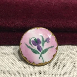 May include: A round brooch featuring a glossy pink enamel surface with a hand-painted purple flower and green leaves at the center. The circular piece is framed by a thin, textured gold-toned metal border with an irregular, scalloped edge.