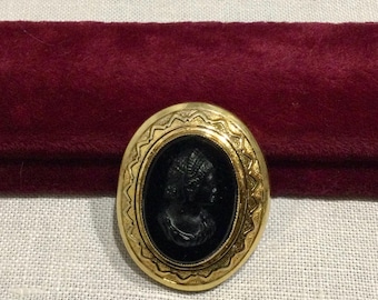 May include: Oval gold-toned brooch with a black cameo of a woman's profile. The brooch has a decorative border and is set against a burgundy velvet background. A classic piece of jewelry.