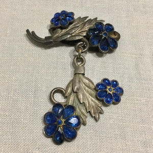 Puede incluir: Broche vintage plateado con diseño floral. Presenta tres grupos de flores azules y un elemento colgante con una hoja y una pequeña flor azul. Las flores tienen un centro dorado.