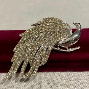 May include: A silver-toned peacock brooch featuring a smooth, polished metal head and neck, with a large, fanned tail intricately detailed with numerous clear, sparkling rhinestones set across the entire plumage surface.