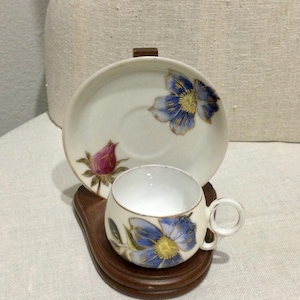 以下が含まれることがあります： 花柄のヴィンテージティーカップとソーサーのセット。白い磁器のカップとソーサーには、金色のトリムが付いた青と黄色の花が描かれています。セットは濃い茶色の木製スタンドに置かれています。