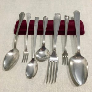 Op de afbeelding: Een verzameling vintage zilverwerk, waaronder lepels en vorken, gerangschikt op een bordeauxrode fluwelen ondergrond. Het zilverwerk heeft een zilveren afwerking en een klassiek ontwerp, perfect voor een formeel diner.