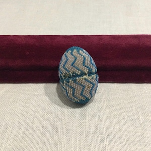 Puede incluir: Un objeto ovalado con cuentas con un patrón de zigzag azul y blanco. El objeto, probablemente un cierre o elemento decorativo, está hecho de pequeñas cuentas. Se encuentra sobre un fondo de terciopelo burdeos y una superficie de lino blanco.
