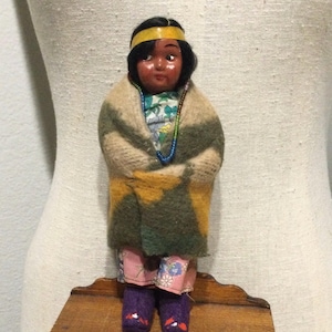Puede incluir: Una muñeca vintage con cabello oscuro, una diadema amarilla y una cara pintada. La muñeca está envuelta en una manta con rayas verdes, amarillas y beige. Lleva una camisa con estampado floral, pantalones rosas y botas moradas. Un collar de cuentas está alrededor de su cuello.