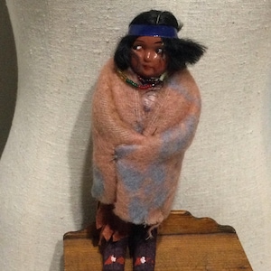 May include: A vintage doll with a painted face, black hair, and a blue headband. The doll is wrapped in a pink and blue patterned blanket. It wears a beaded necklace and dark purple boots with red and white accents. The doll is standing on a wooden surface.