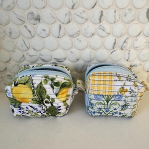 May include: Two small, quilted fabric pouches with blue zippers. One features a lemon and lavender print, the other a yellow plaid and floral design. Both have a small fabric loop on the side.